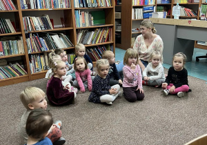 Krasnale siedzą na dywanie z Wychowawcą w bibliotece i słuchają Pani czytającej im bajkę.
