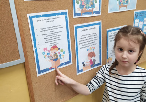 Dziewczynka stoi przed tablicą z obrazkami dotyczącymi bezpiecznego korzystania z Internetu. Wskazuje obrazek z wymienionym korzyściami korzystania z Internetu.