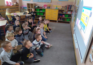 Dzieci siedzą na dywanie przez tablicą interaktywną i oglądają film edukacyjny pt. „Szanuj innych w sieci.”