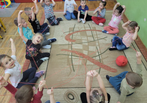 Dzieci z grupy Stokrotki tworzyły własną sieć internetową. Dzieci siedzą dookoła dywanu i trzymają w rączkach włóczkę przeplataną w różne strony.