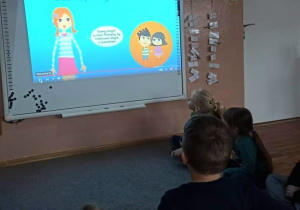 Dzieci z grupy Smerfy siedzą na dywanie. W tle tablica interaktywna, na której wyświetlany jest film edukacyjny "Szanuj innych w sieci".