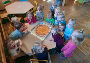 Dzieci z grupy Krasnale stoją wokół stołu, na którym znajduje się pizza.