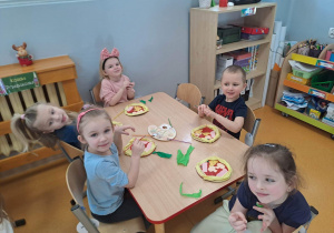 Dzieci siedzą przy stoliku i wykonują papierową pizzę z gotowych elementów.