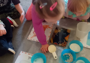 Na dywanie ustawione są w pojemnikach: mąka, cukier, sól, drożdże. Dziewczyna w różowej bluzce wspólnie z nauczycielką odczytuje wyraz i łączy pojemnik z obrazkiem.