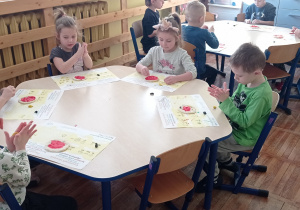 Przy stoliczku siedzi pięcioro dzieci. Przed przedszkolakami leżą uformowane z masy solnej pizze, które zostały pomalowane czerwoną farbą. Dzieci zajmują się lepieniem z plasteliny składników do pizzy.