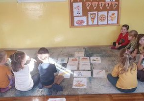 Dzieci siedzą na dywanie przed tablicą z napisem Dzień Pizzy.