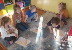 Przedszkolaki siedzą na dywanie przed sobą mają obrazki na których narysowany jest trójkąt, przedstawiający kawałek pizzy. Wybierają karteczki z obrazkami przedstawiającymi składniki na pizze i układają je na swoich kartkach.
