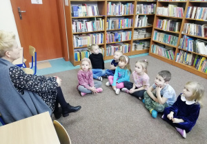 Przedszkolaki siedzą na dywanie i słuchają informacji podawanych przez Panią.