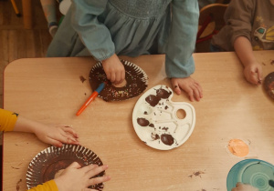 Dzieci przy stolikach dekorują pączki z tacek papierowych, wcześniej pomalowany na brązowo posypkami.