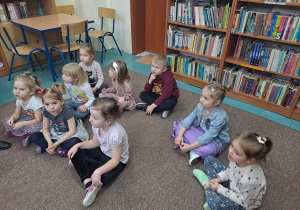 Na zdjęciu widać dzieci, które z zaciekawieniem słuchają pani pracującej w bibliotece.