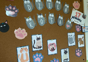 Zdjęcie dekoracji na tablicy z okazji Dnia Kota. Wiszą literki tworzące napis oraz tematyczne grafiki przedstawiające koty.