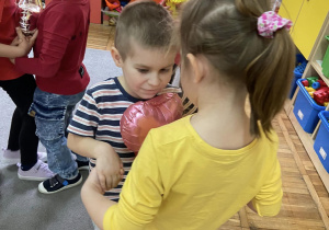 Dzieci tańczą w parach, trzymając się za ręce i starają się utrzymać balon w kształcie serca swoimi brzuszkami.