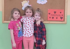 Trzy dziewczynki obejmują się na tle tablicy z walentynkową dekoracją