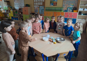Dzieci stoją przy stoliku na którym leżą produkty spożywcze przeznaczone do pieczenia pączków.
