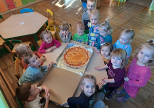 Dzieci stoją szczęśliwe przy stoliczku na którym leży świeża i gorąca pizza.