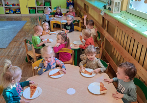 Dzieci siedzą przy stoliczkach i zajadają apetycznie pizzę.