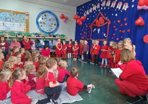 Dzieci ubrane na czerwono siedzą w holu przedszkolnym i oglądają występ w wykonaniu Stokrotek.