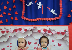 Chłopiec i dziewczynka pozują w fotobudce wystawiając uśmiechnięte twarze z wyciętych otworków planszy związanej z Walentynkami.