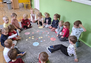 Dzieci siedzą w kole na dywanie i wykonują ćwiczenie w dopasowaniu pączka w odpowiednim kolorze do talerzyka w tym samym kolorze.