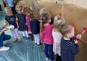 Dzieci z pędzlami w dłoni tworzą dzieło malarskie na ogromnym papierze przyklejonym do szafy.