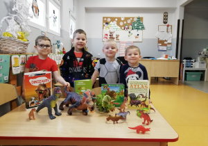 Chłopcy stoją za ławką, na której stoją plastikowe dinozaury oraz książki o ich tematyce.