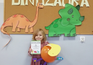 Dziewczynka stoi na tle tablicy z napisem Dzień Dinozaura i trzyma w ręku dyplom oraz dinozaura wykonanego z balonu.
