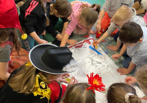 Na zdjęciu znajdują się dzieci w trakcie warsztatów, które siedzą na podłodze i malują farbami obrazek.