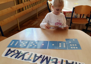 Dziewczynka siedzi przy stoliku, układa domino z kropkami. Na stoliku leży napis matematyka.
