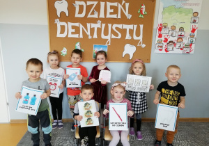 Przedszkolaki stoją na tle tablicy z napisem Dzień Dentysty i w rękach trzymają obrazki rzeczy związanych z higieną jamy ustnej.