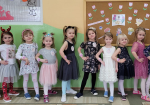 Wszystkie dziewczynki z grupy Misie stoją bokiem, z rączką na bioderku pod tablicą-prezentacja kandydatek na Małą Miss