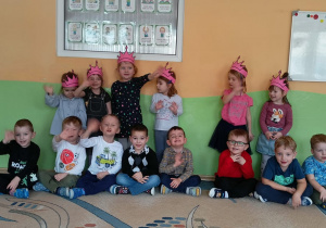 Dziewczynki z grupy Biedronki stoją w różowych koronach, chłopcy siedzą i machają.