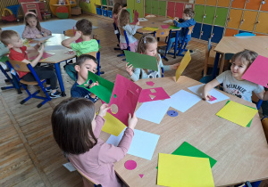 Dzieci siedzą przy stolikach i wypychają figury geometryczne z kart pracy.