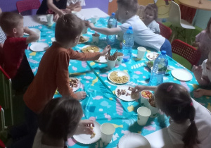Zdjęcie przedstawia grupę przedszkolaków siedzących przy stole i częstujących się słodyczami.