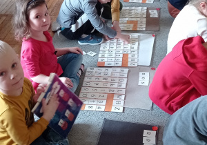 Grupa przedszkolaków siedzi na dywanie, przed sobą mają maty i klasery z literami. Dzieci układają z kartonowych tabliczek napis : „nos”.