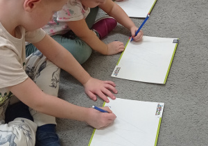 Grupa dzieci siedzi na dywanie, każde dziecko ma przed sobą małą tablicę zmywalną. Przedszkolaki próbują napisać po śladzie literkę „M”.