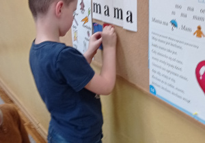 Chłopiec ubrany na niebiesko stoi przy tablicy do której przyczepia czerwony kartonik.