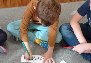 Chłopiec siedzi na dywanie. Przed sobą ma kartkę z napisem swoje imienia na której zaznacza nową poznaną literkę „M”.