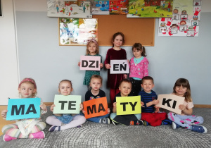 Dzieci siedzą na dywanie i w rękach trzymają kartki z literami, z których układa się napis Dzień Matematyki.