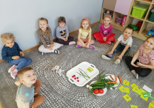 Dzieci siedzą dookoła tacy, na której leży działanie matematyczne – dodawanie ułożone z warzyw.