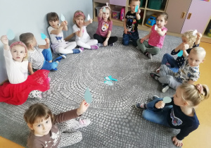 Przedszkolaki siedzą na dywanie i pokazują trzymane w dłoniach papierowe krople.