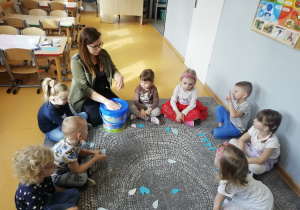 Nauczycielka uderza w plastikowe pudełko, w kole siedzą dzieci przed, którymi lezą papierowe krople