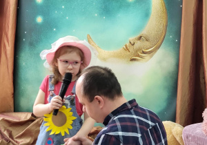 Dziewczynka śpiewa do mikrofonu. Ubrana jest w dżinsową sukienkę przyozdobioną kwiatami, na głowie ma kolorowy kapelusz, a w ręku koszyczek. Przed nią klęczy tata, który trzyma ją za rękę.