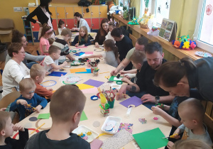 Dzieci z rodzicami siedzą wspólnie przy stolikach i robią papierowe świąteczne wianki.