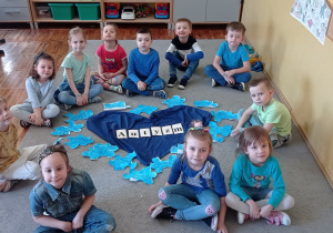 Jedenaścioro dzieci siedzi na dywanie wokół napisu „autyzm” ułożonego na niebieskim materiale w kształcie serca. Wokół dekoracji ułożone są prace dzieci: kartki wycięte w kształcie puzzli z napisami dotyczącymi zachowań osób z autyzmem.
