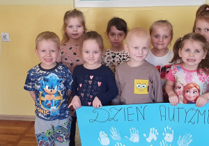 Dzieci z grupy Słoneczka pozują do zdjęcia trzymając plakat z napisem Dzień Autyzmu oraz odbitymi dłońmi na niebieskim tle.