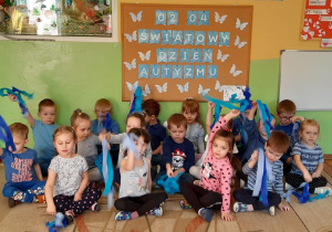 Dzieci z grupy Stokrotki pozują do zdjęcia przy tablicy z napisem 02.04. Światowy Dzień Autyzmu wymachując niebieskimi wstążkami.