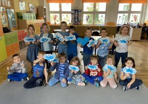 Dzieci z grupy Żabki pozują do zdjęcia. W rękach trzymają motylki, które zostały wykonane poprzez odbicie swoich pomalowanych niebieską farbą dłoni. Jeden chłopiec trzyma niebieskie serce z napisem „Autyzm”.