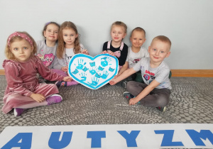 Dzieci z grupy Liski siedzą na dywanie za napisem autyzm i trzymają serce z odbitymi na niebiesko dłońmi oraz doklejonymi niebieskimi motylami.