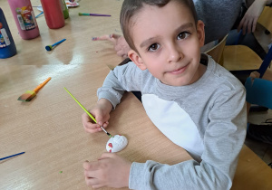 Chłopiec siedzi przy stoliku trzyma w ręku pędzelek i maluje jajeczko z gipsu. Na stole leżą pędzelki, farby. Obok siedzą inne dzieci.