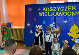 Dziewczynka w stroju owieczki oraz chłopiec stoją obok siebie i recytują wierszyki, reszta dzieci siedzi z tyłu na dywanie.